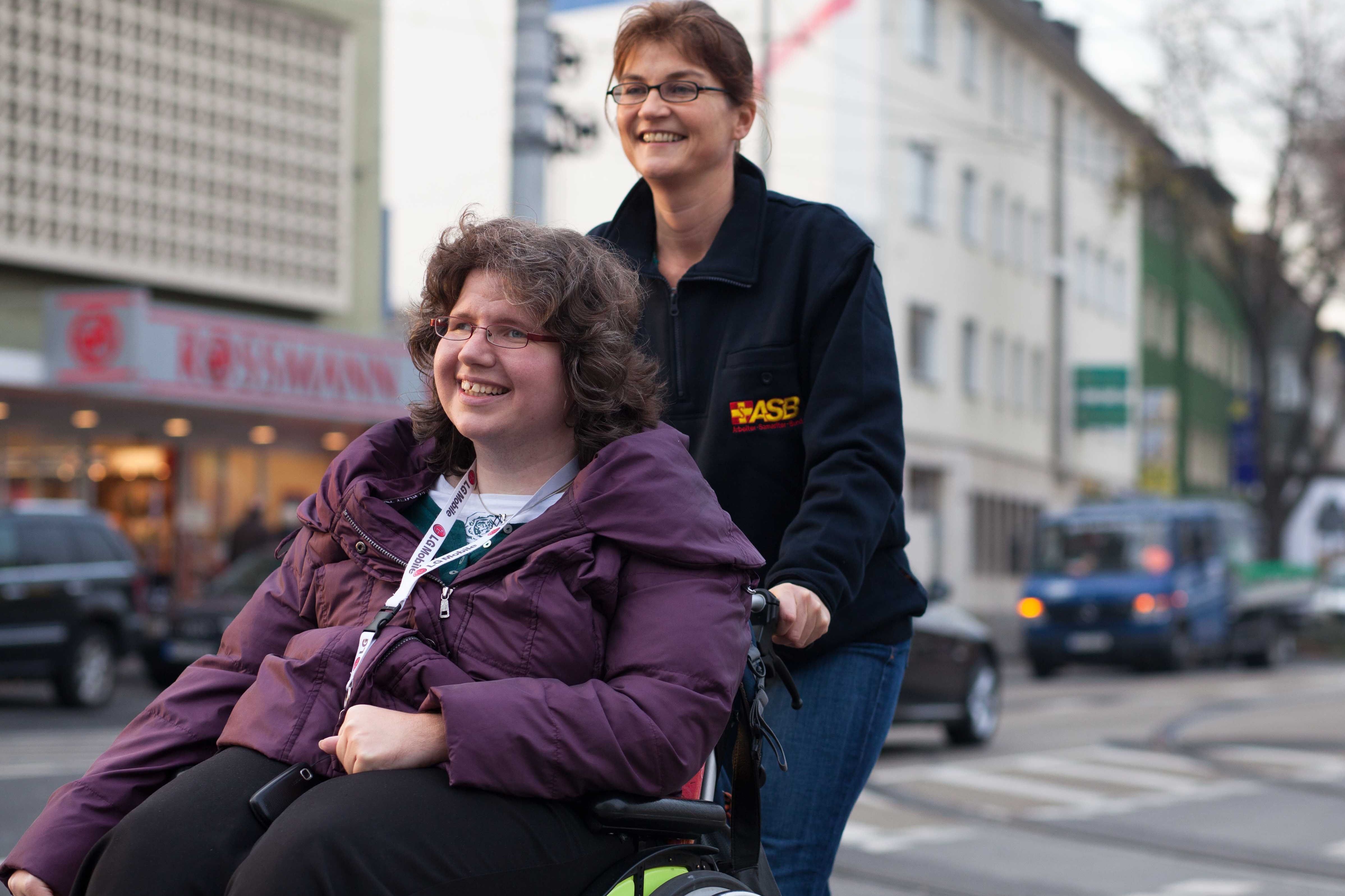 Individuelle Schwerstbehindertenbetreuung Eine Frau schiebt eine andere Frau im Rollstuhl im Straßenverkehr