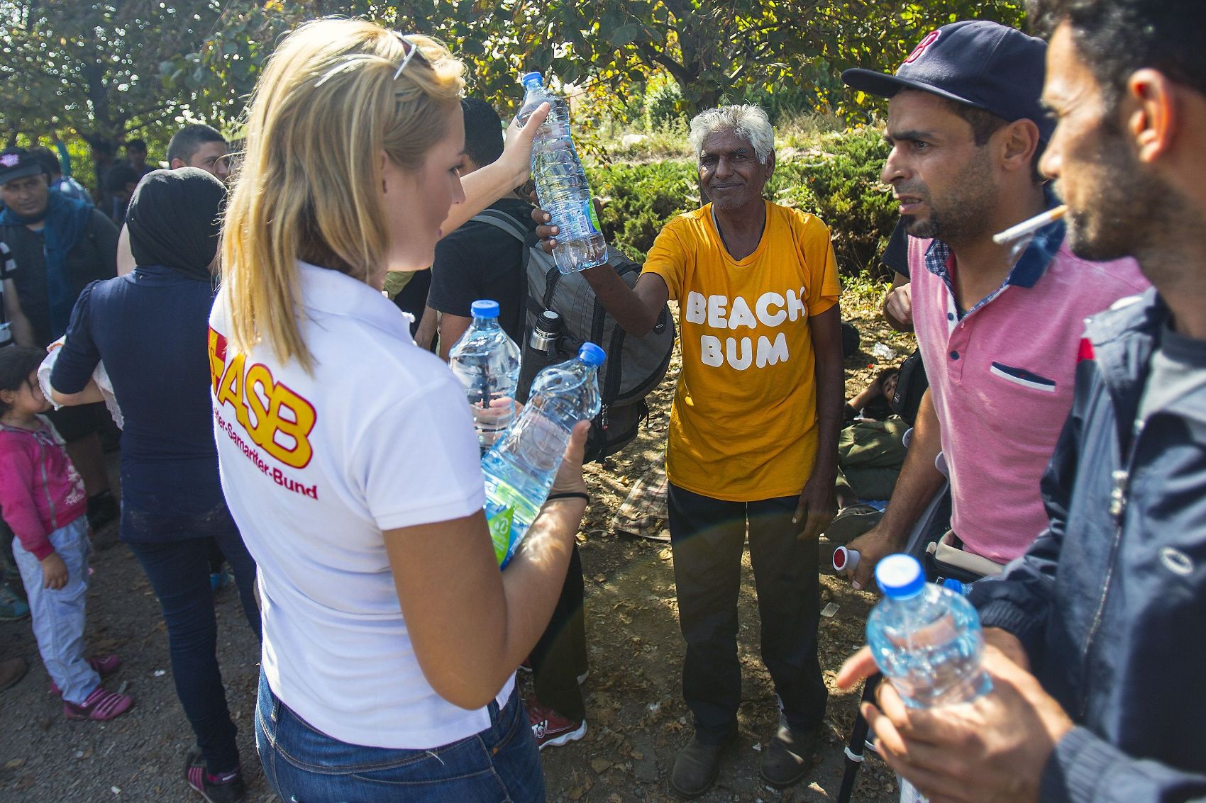 Eine ASB-Helferin verteilt Wasserflaschen an Flüchtlinge, die im Schatten am Straßenrand warten.